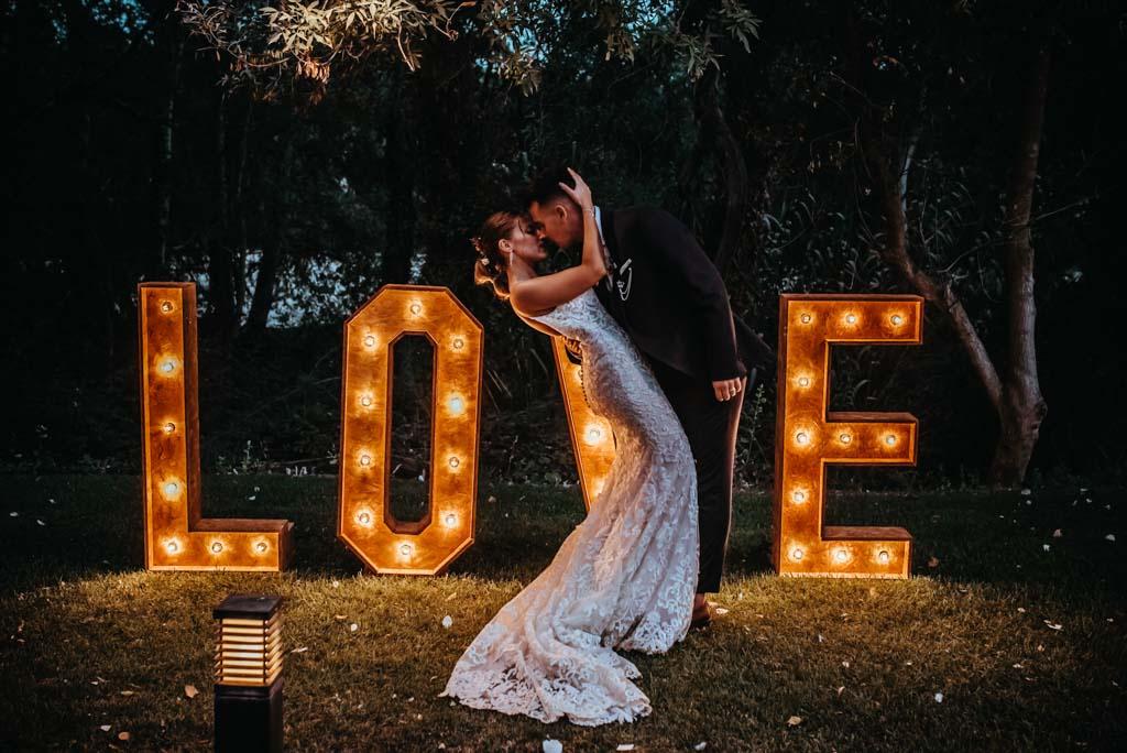 Pareja besándose el día de la boda frente a unas letras iluminadas en las que puede leerse la palabra Love