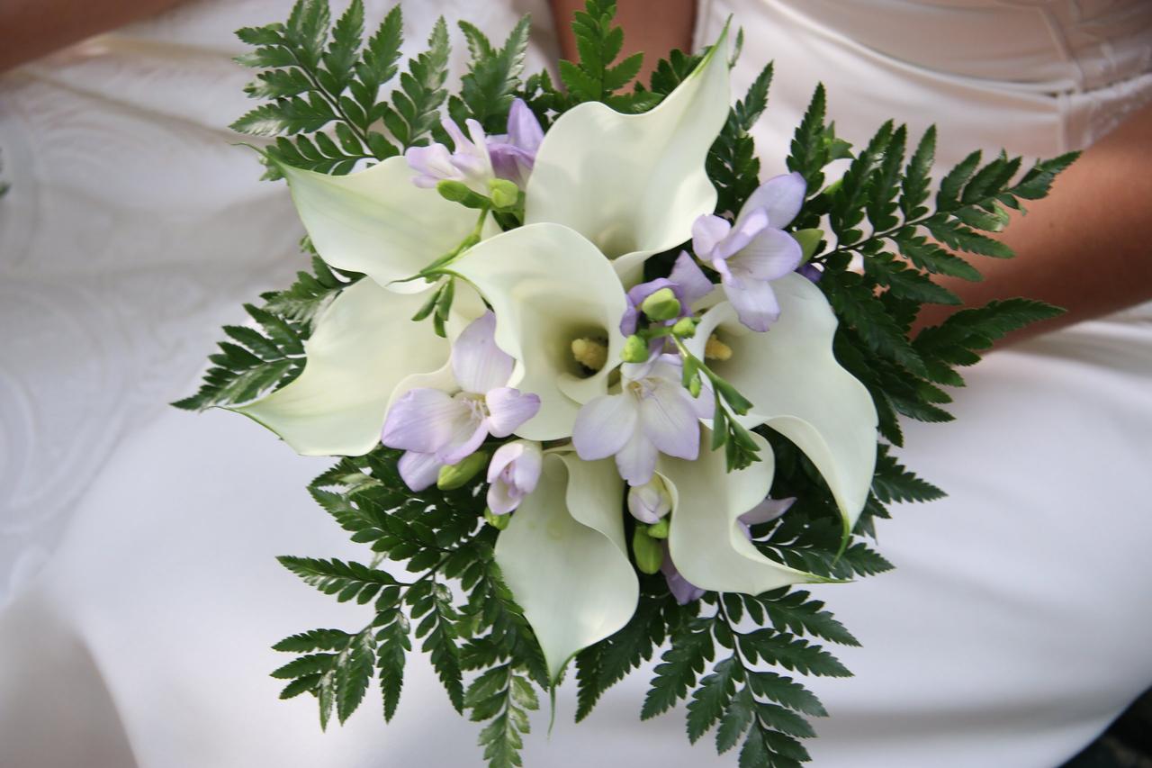 Chica sentada sujeta en sus manos un ramo de novia con calas blancas, mucho verde y unas flores lilas