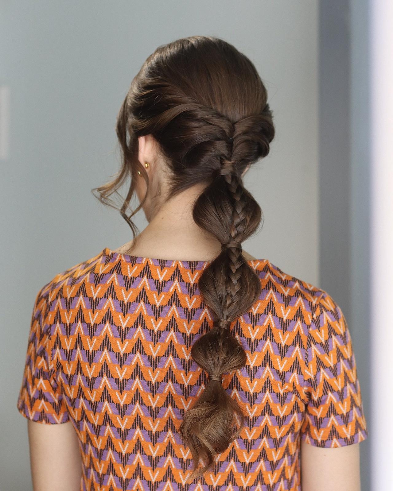 peinados trenzas boda: mujer de espaldas con vestido estampado naranja y pelo moreno con coleta de burbujas decorada con con trenza por encima