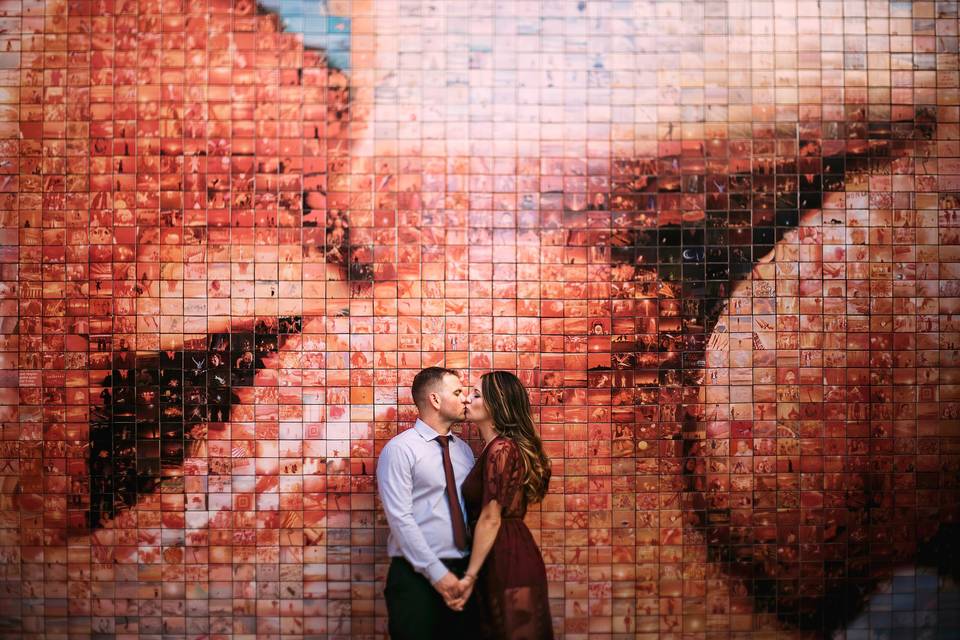 Pareja con las manos cogidas se besa frente a un muro con la imagen de dos bocas besándose