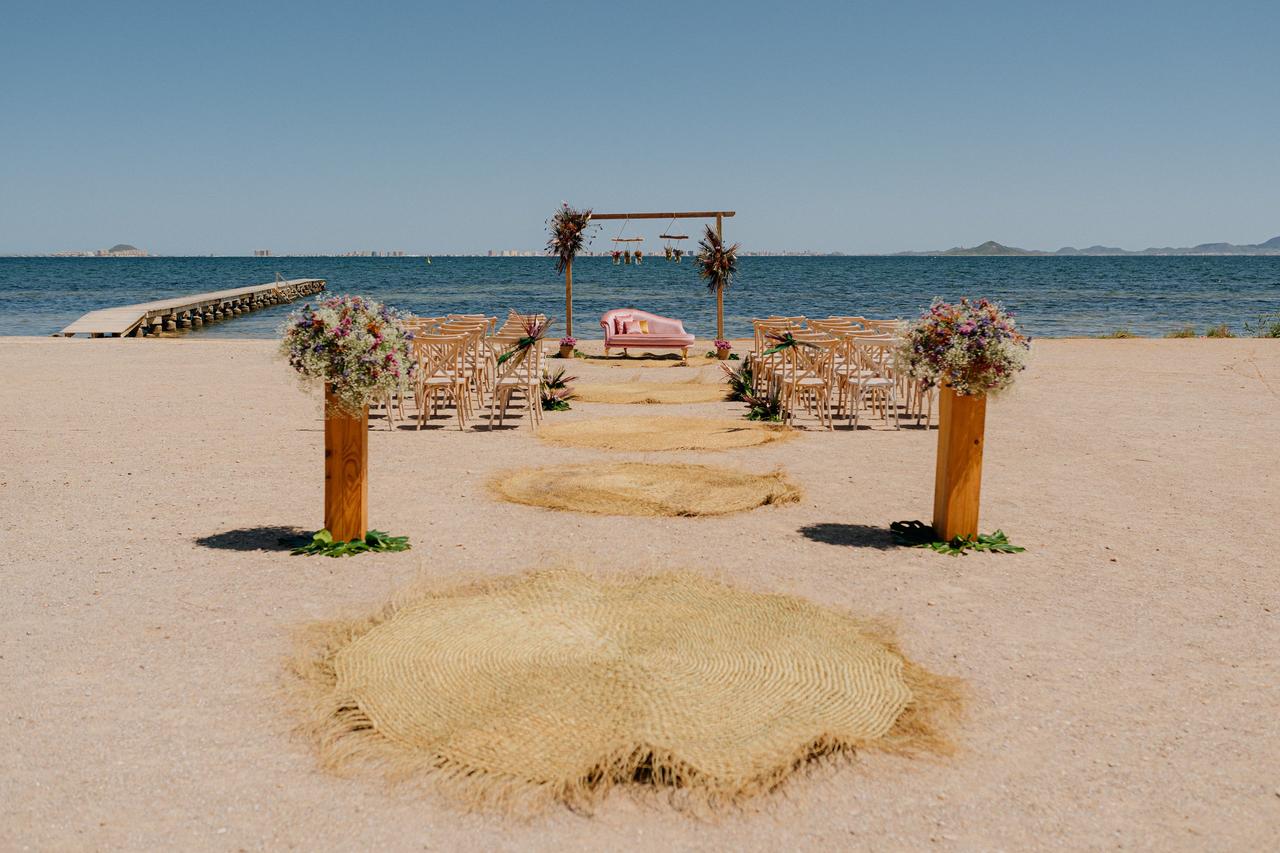 Bonita playa con todo a punto para celebrar una boda civil al aire libre con maceteros, alfombras, sillas de madera, un altar y un sofá rosa