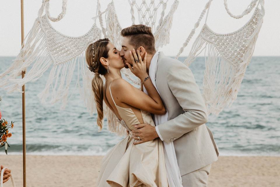 Chico y chica vestidos de novios y besándose mientras se abrazan en el altar de su boda civil en la playa