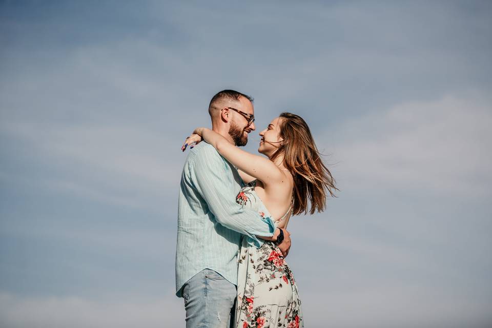 pareja de chico y chica mirándose a los ojos abrazados con el cielo de fondo