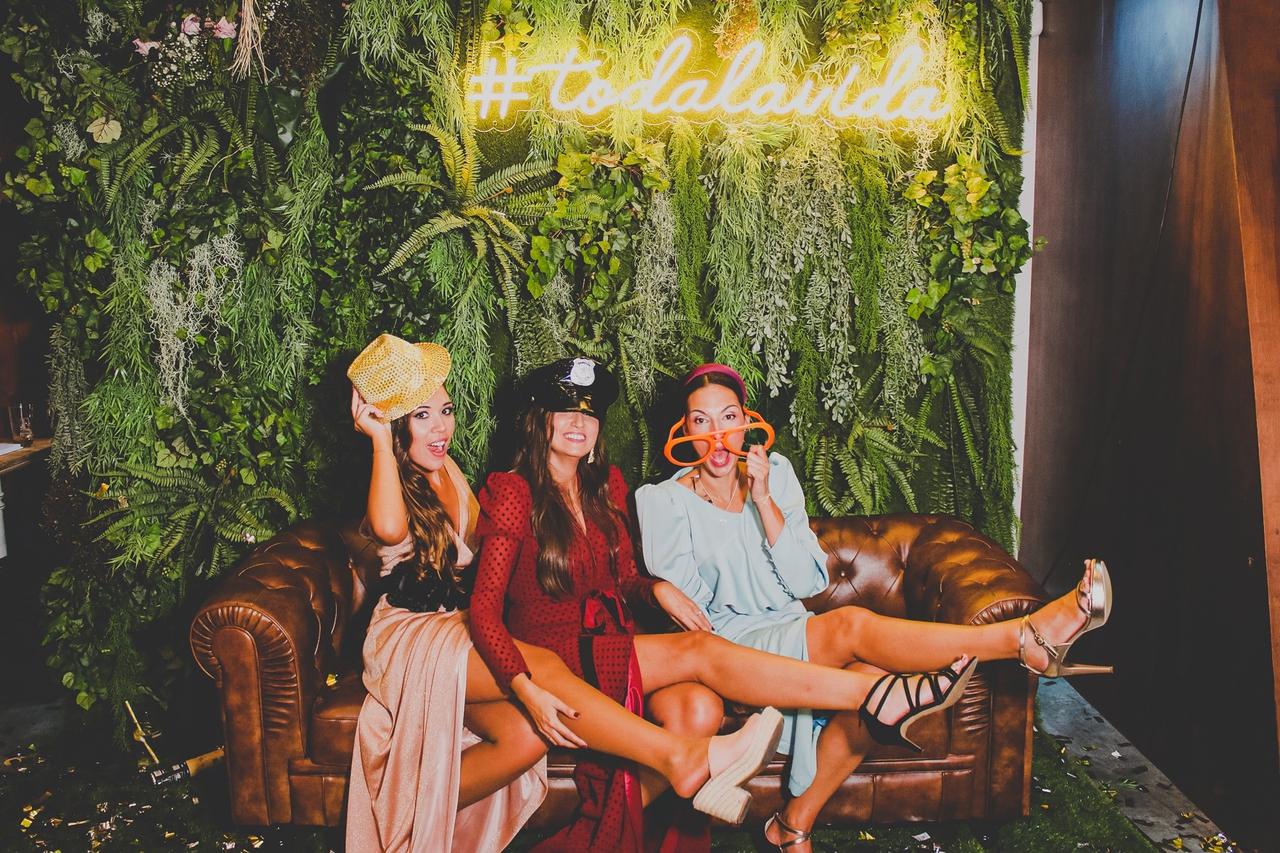 Tres chicas con vestidos de fiesta y un divertido atrezo sentadas en un sofá de tipo chester bajo un neón luminoso con el hashtag 