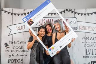 Tres invitadas posan sonrientes en un photocall de boda con un marco como los de Facebook