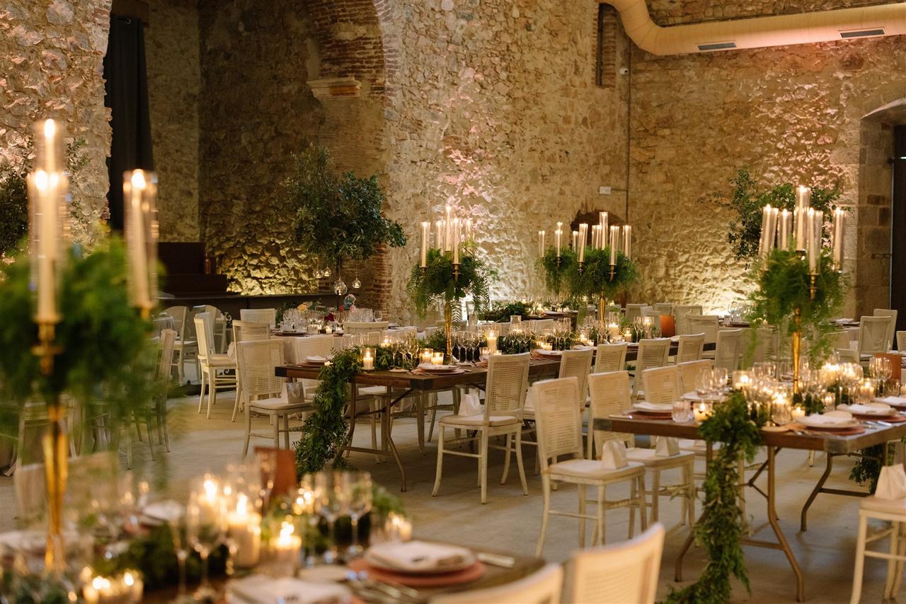 Precioso espacio con paredes de piedra y varias mesas rectangulares preparadas para un banquete de boda