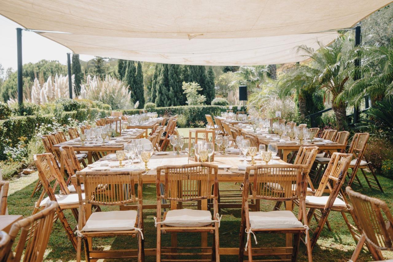 Jardín con un toldo que cubre varias mesas rectangulares de madera y diferente capacidad, bien vestidas para una comida o cena