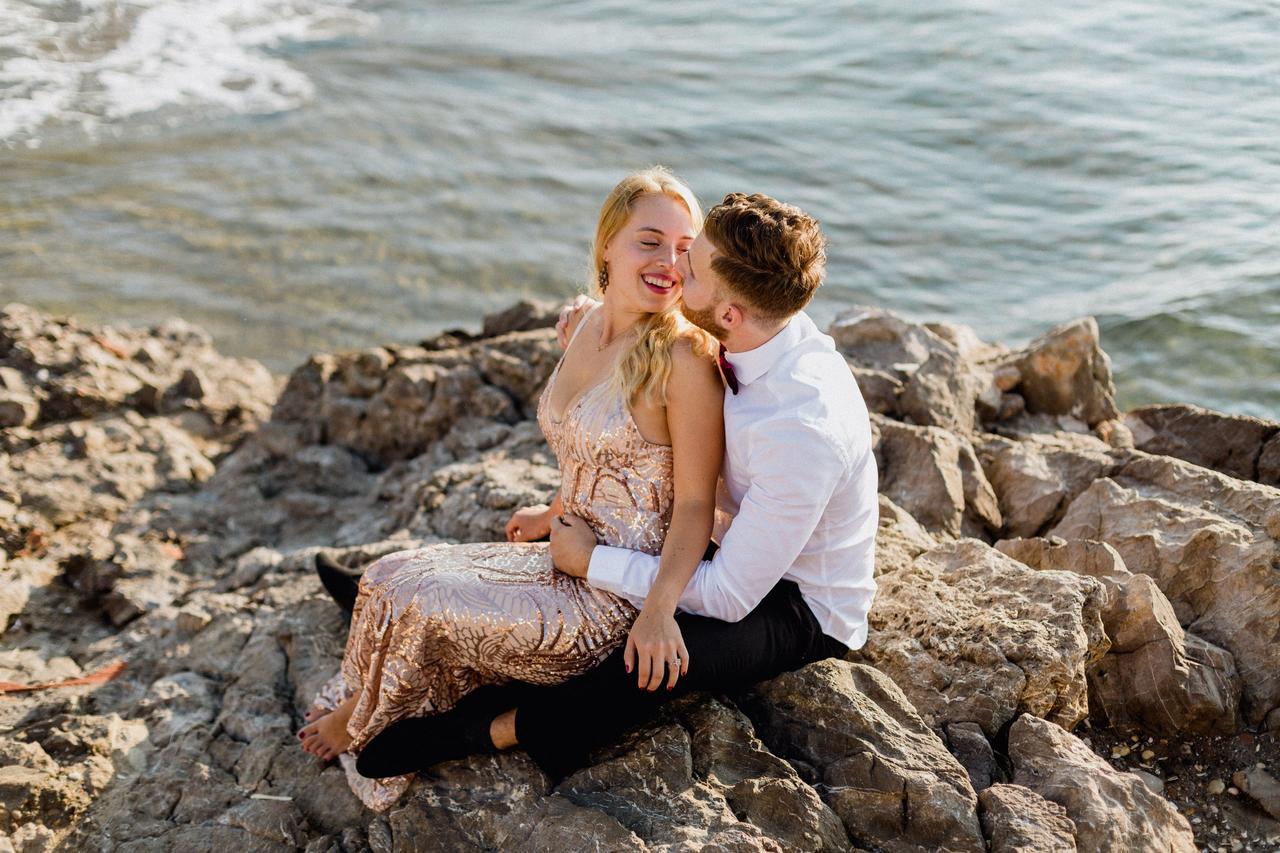 Chica rubia con un vestido dorado sentada encima de las piernas de un chico sobre unas rocas, junto al mar