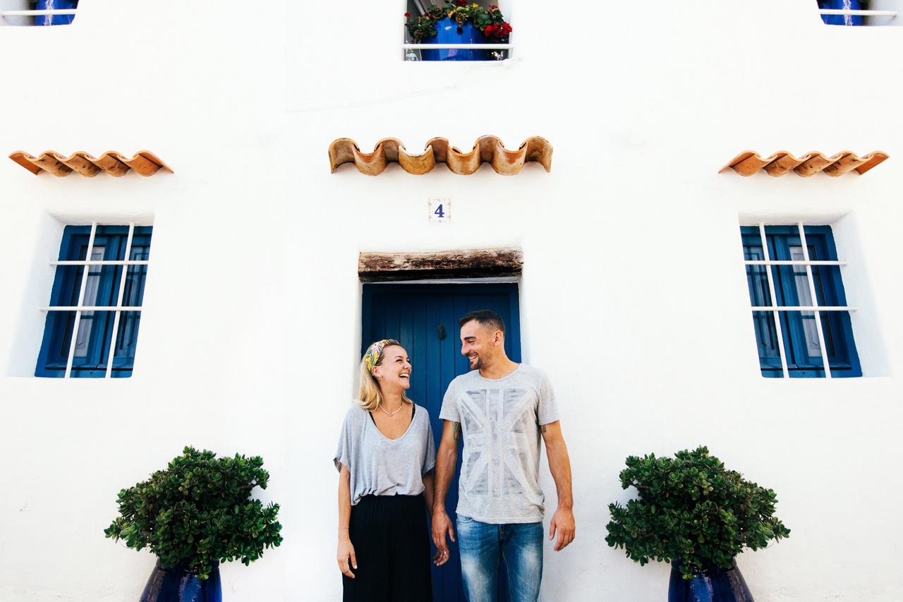 Chico y chica vestidos de forma informal se miran muy sonrientes delante de una casa con una puerta en color azul