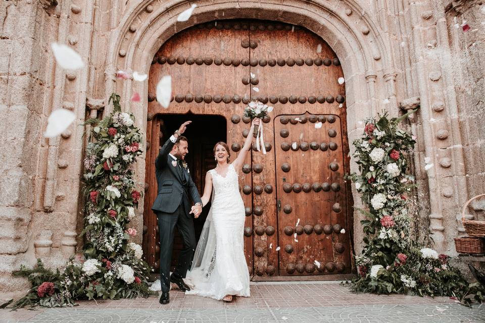 Pareja de recién casados saliendo de la iglesia felices y cogidos de la mano mientras reciben una lluvia de arroz y pétalos de flores 