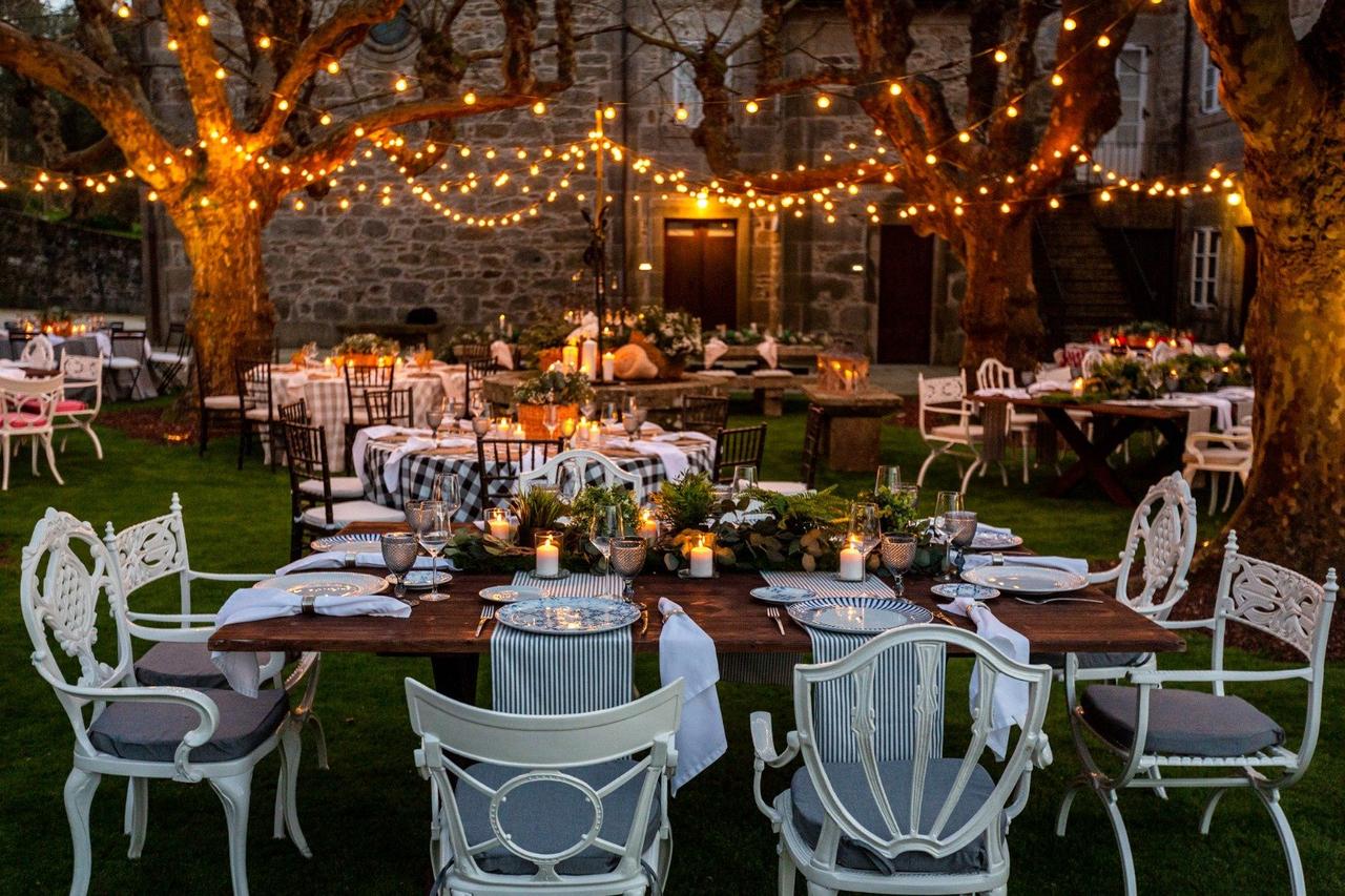 Jardín al atardecer con mesas y sillas de diferentes modelos preparadas para una cena, perfectamente iluminadas con guirnaldas de luces