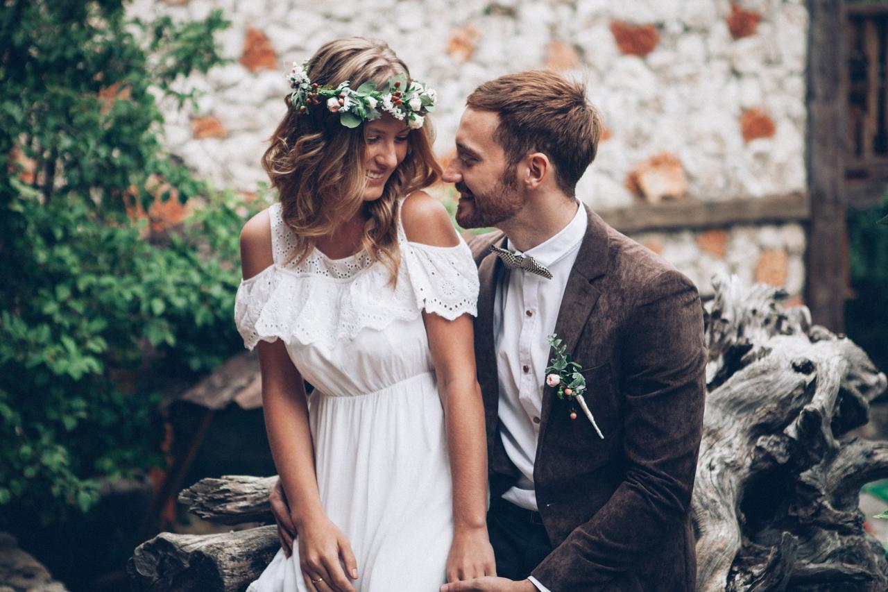 Invitar a un ex a la boda: chica vestida de novia muy sonriente sentada en la falda de un chico con traje marrón en un exterior