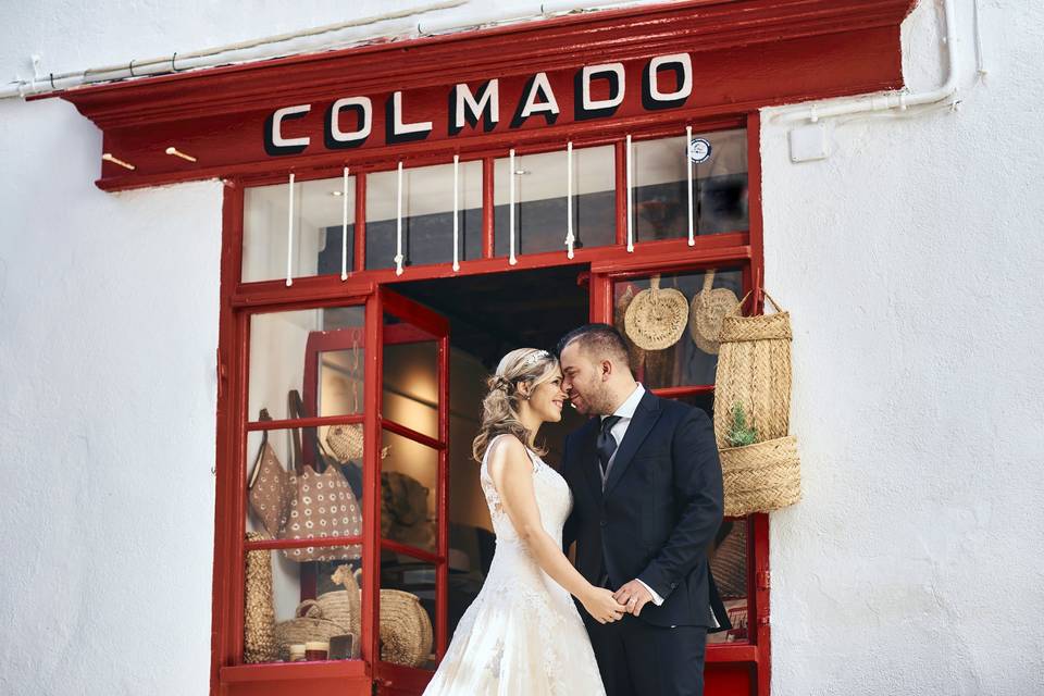 Cuánto cuesta una boda: pareja vestida de novio y novia muy juntos y cogidos de la mano delante de un colmado