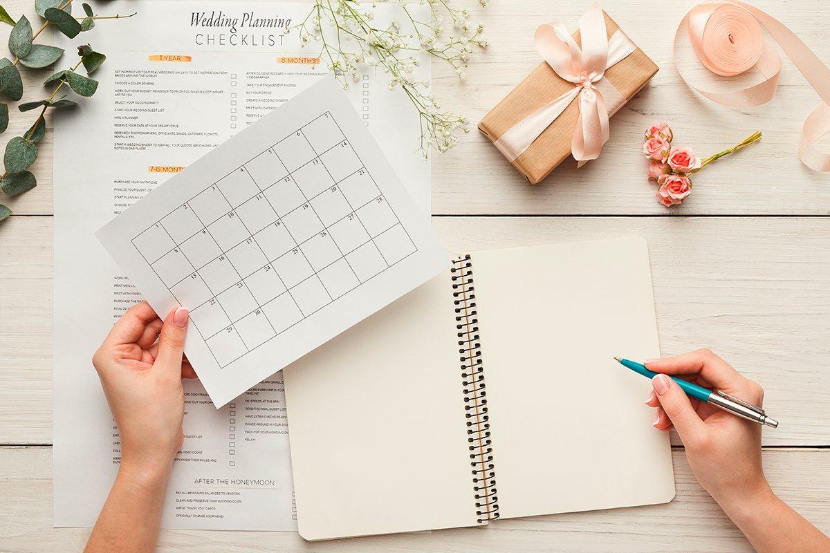 Mesa con una libreta y una mano femenina que sostiene por un lado un bolígrafo y por otro una hoja con un calendario en blanco