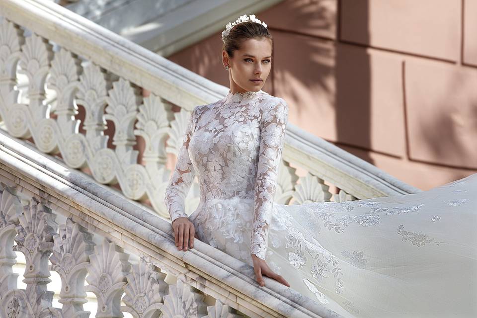 Chica con los ojos claros y el pelo recogido hacia atrás, posa en una escalera con un vestido de novia de manga larga con motivos florales y llevando una diadema en la cabeza