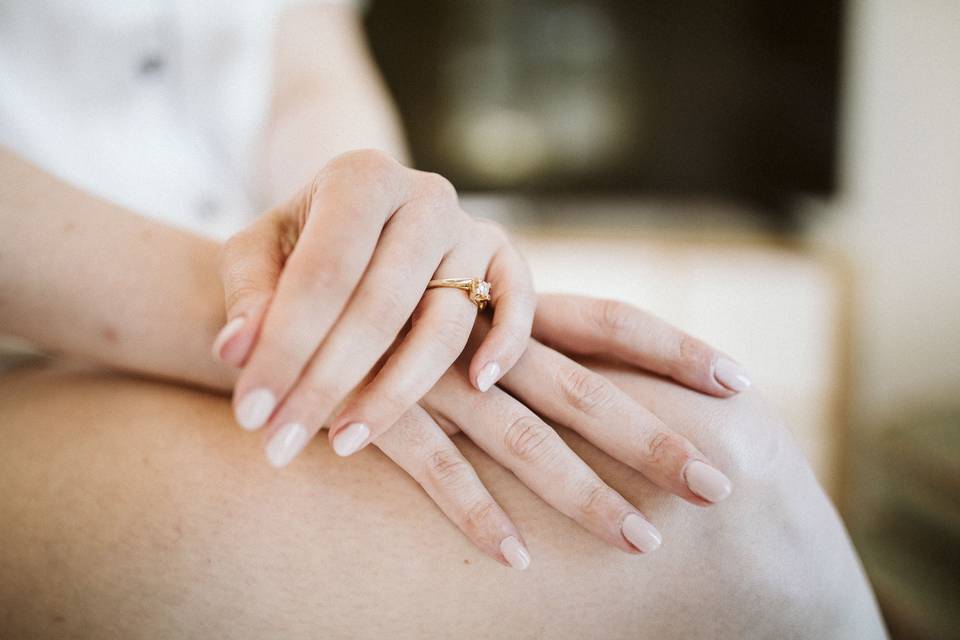 Manos femeninas cruzadas sobre una pierna con la manicura en rosa nude y un anillo de compromiso en oro amarillo
