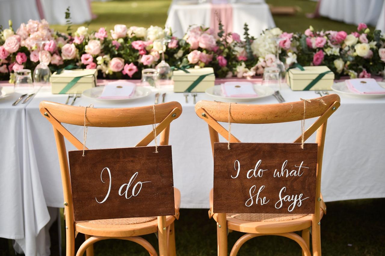 Sillas de los novios el día de la boda con carteles en los que se lee I do y I do what she says