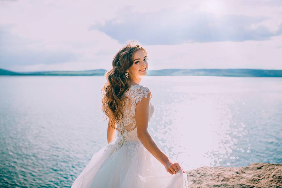 Chica sonriente junto al mar con el pelo suelto y un precioso vestido de novia con encaje