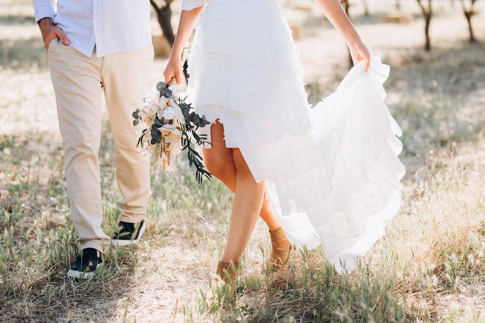 Piernas de chico con pantalón beige y de chica con vestido blanco y un ramo de flores en la mano mientras caminan por medio de la naturaleza