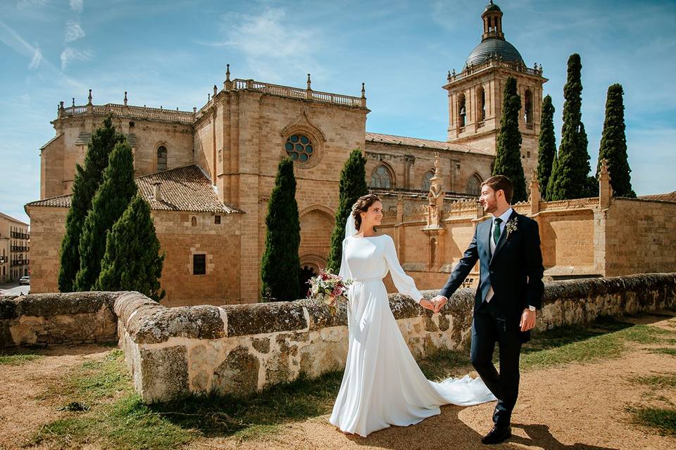 Pareja vestida de novio y novia caminan de la mano sonrientes frente a la catedral de Ciudad Rodrigo