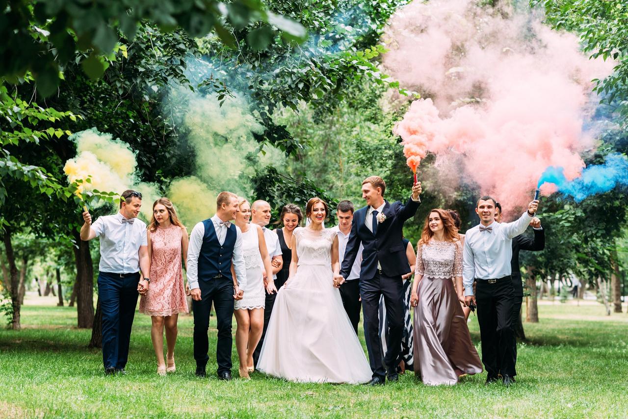 Invitar a un ex a la boda: pareja de recién casados pasean cogidos de la mano por un jardín con amigos y bengalas de humo de colores