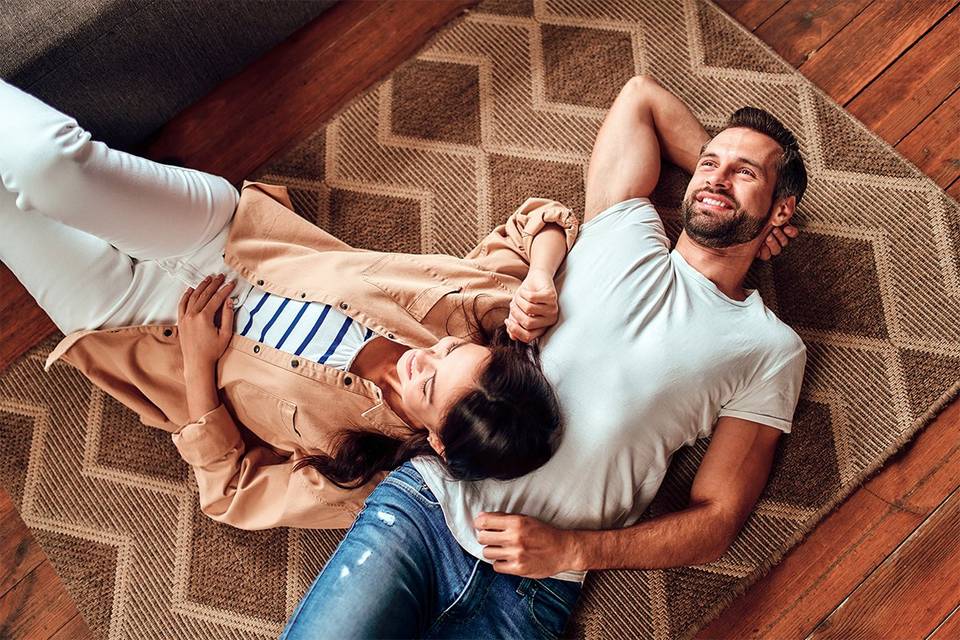 Pareja de chico y chica feliz estirada en una alfombra con ropa informal sobre un suelo de madera