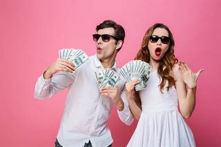 Retrato de una pareja alegre y elegantemente vestida sosteniendo un montón de billetes de dinero y celebrando aislados sobre fondo rosa