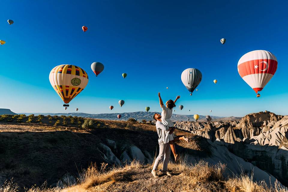 Planes para hacer en pareja: chico y chica felices en un entorno natural con globos aerostáticos volando por encima de ellos