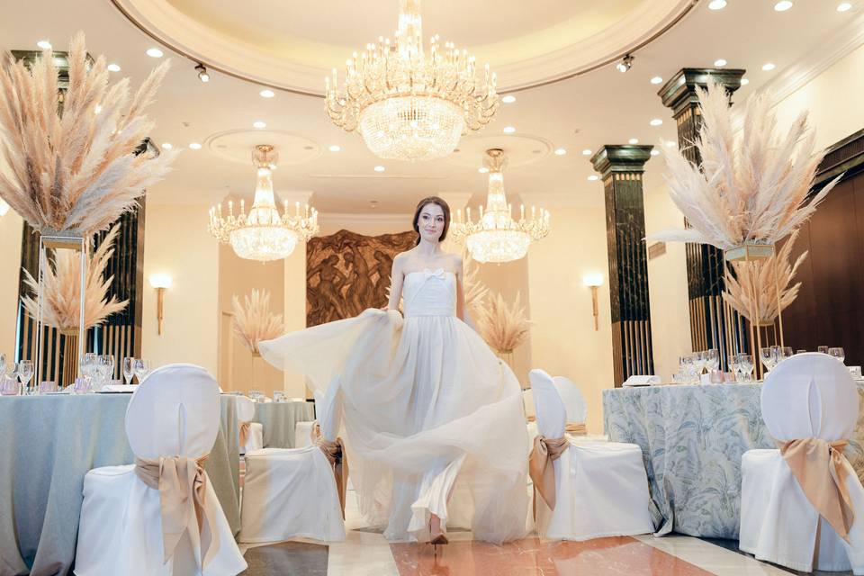 Chica vestida de blanco pasea sonriente en un salón con varias mesas elegantemente vestidas para una comida o cena especial