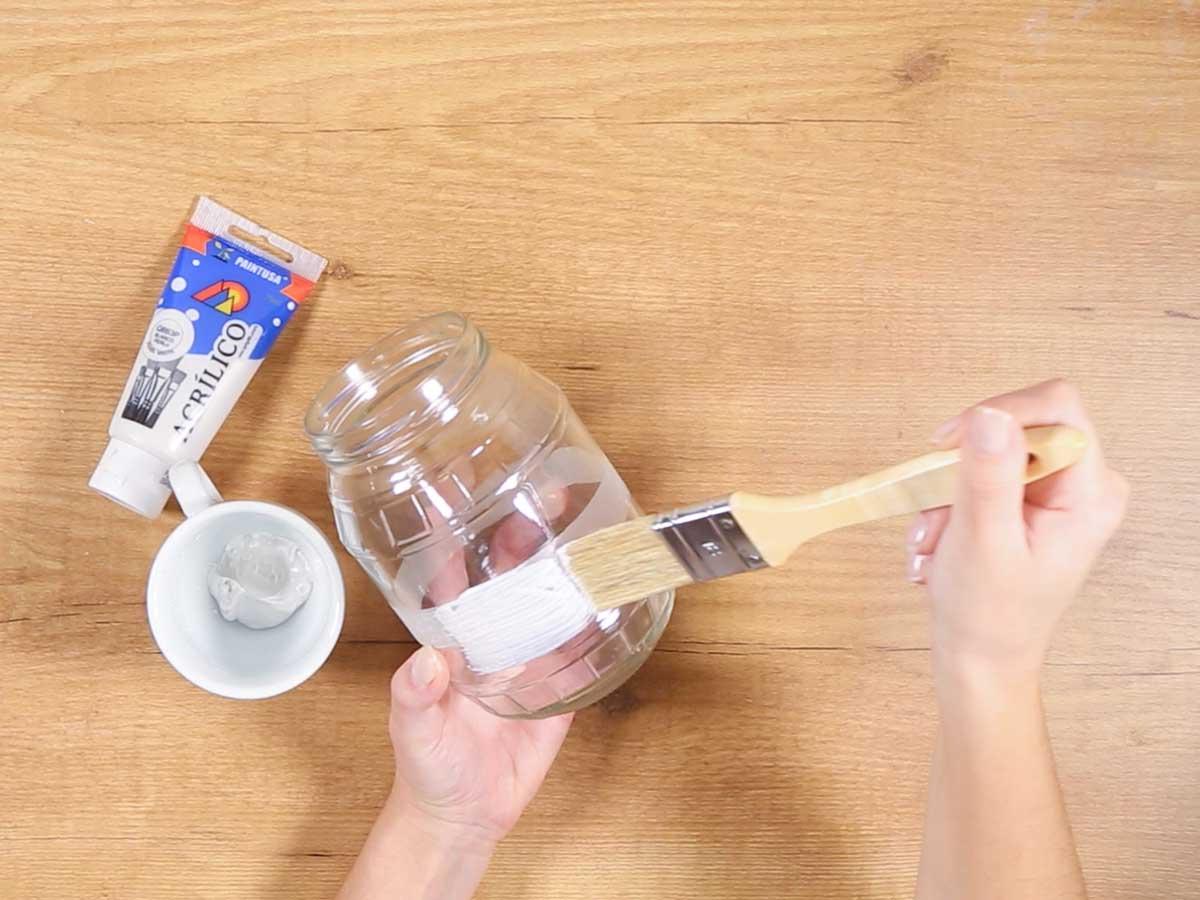 Manos pintando de blanco un tarro de cristal transparente con la ayuda de un pincel