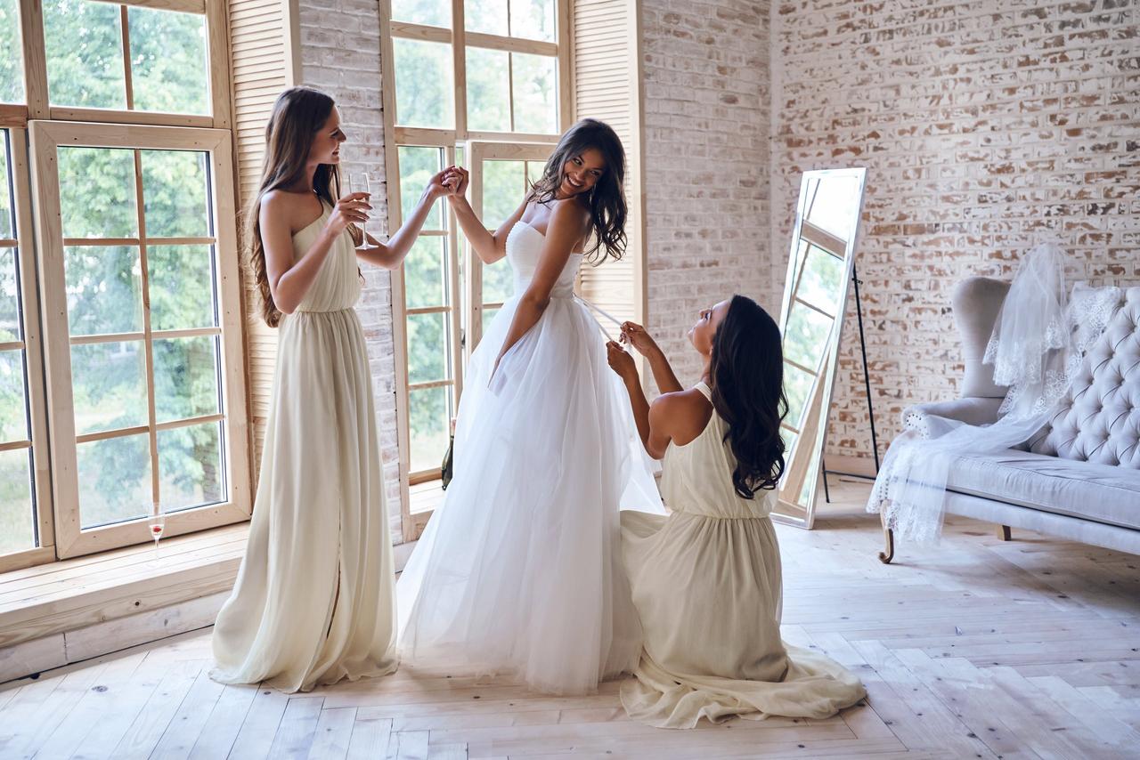 Cosas que las novias olvidan hacer el día de su boda: novia sonriente mientras una amiga le ayuda a acabar de ponerse el vestido de novia y otra le coge la mano