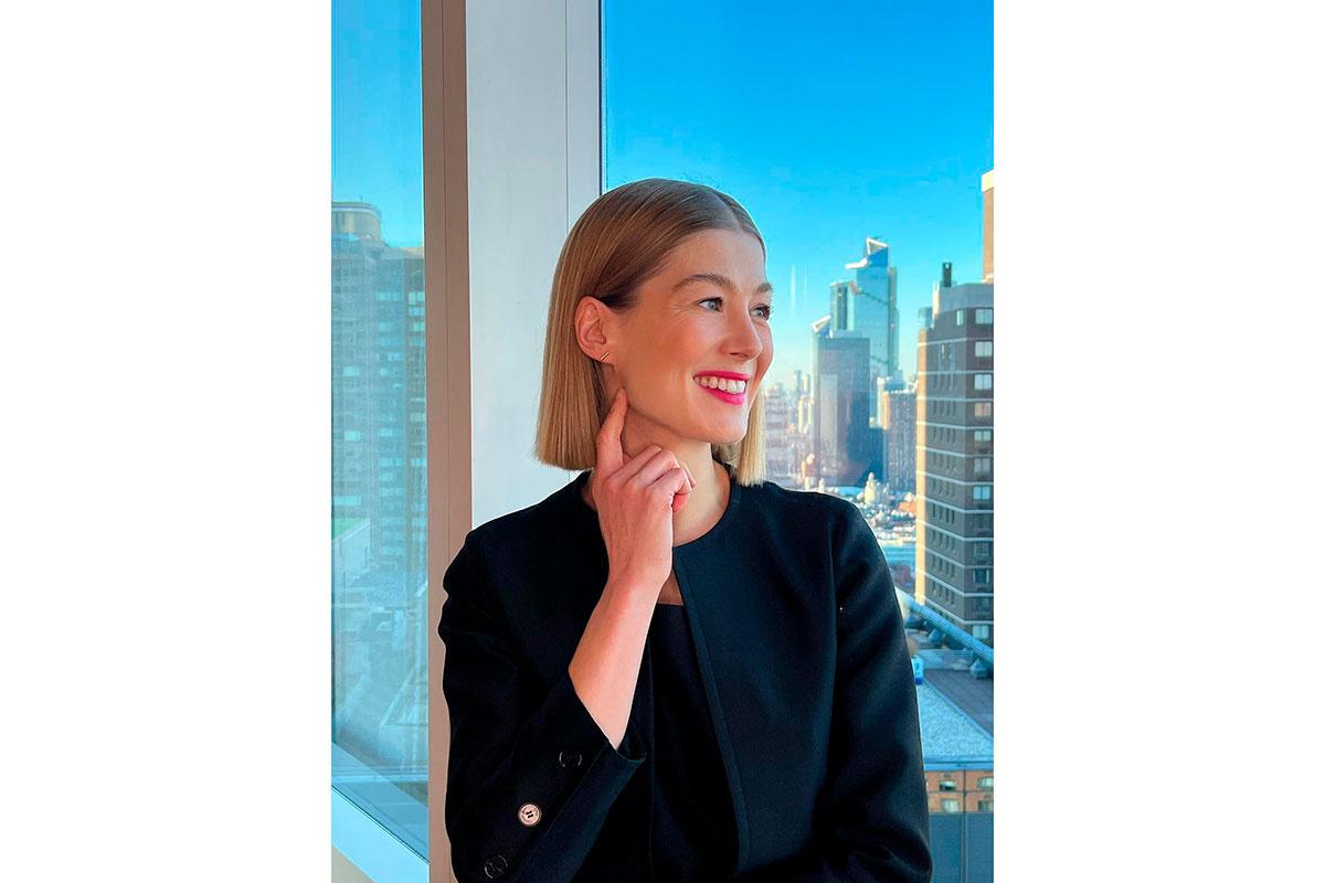 Rosamund Pike con melena corta pulida con un paisaje de edificios altos de fondo