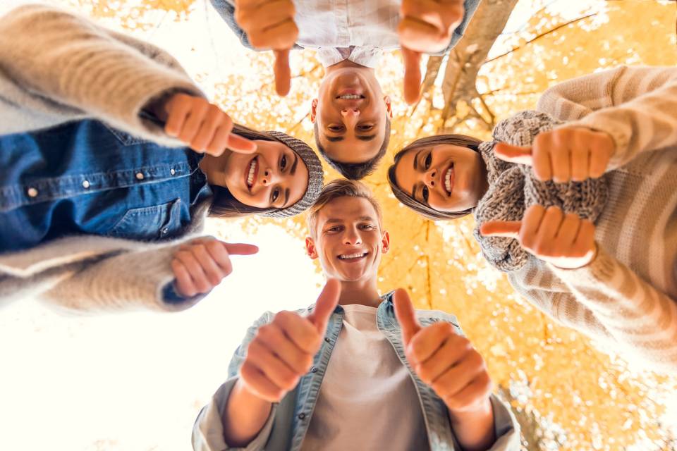 Fotografía desde el suelo de 4 chicos y chicas jóvenes y sonrientes con los pulgares de los dedos hacia arriba
