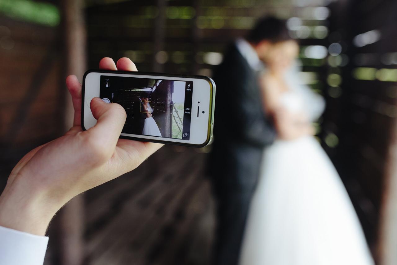 Wedding content creator: teléfono móvil y mano en primer plano grabando a una pareja de recién casados vestidos con traje clásico y vestido de novia