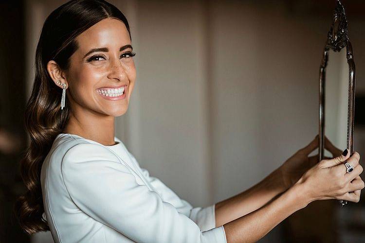 María F. Rubíes sonriente con pelo ondulado mirando a cámara y sujetando un espejo con las manos