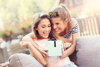 Una chica joven sorprende a otra sentada en un sofá con un regalo en una caja cuadrada