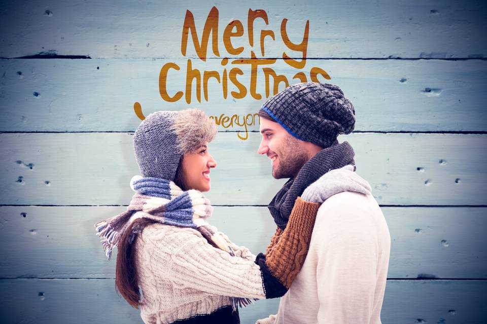 Compromiso en Navidad: pareja de chico y chica abrazados, mirándose a los ojos y muy abrigados delante de un letrero en el que pone Merry Christmas