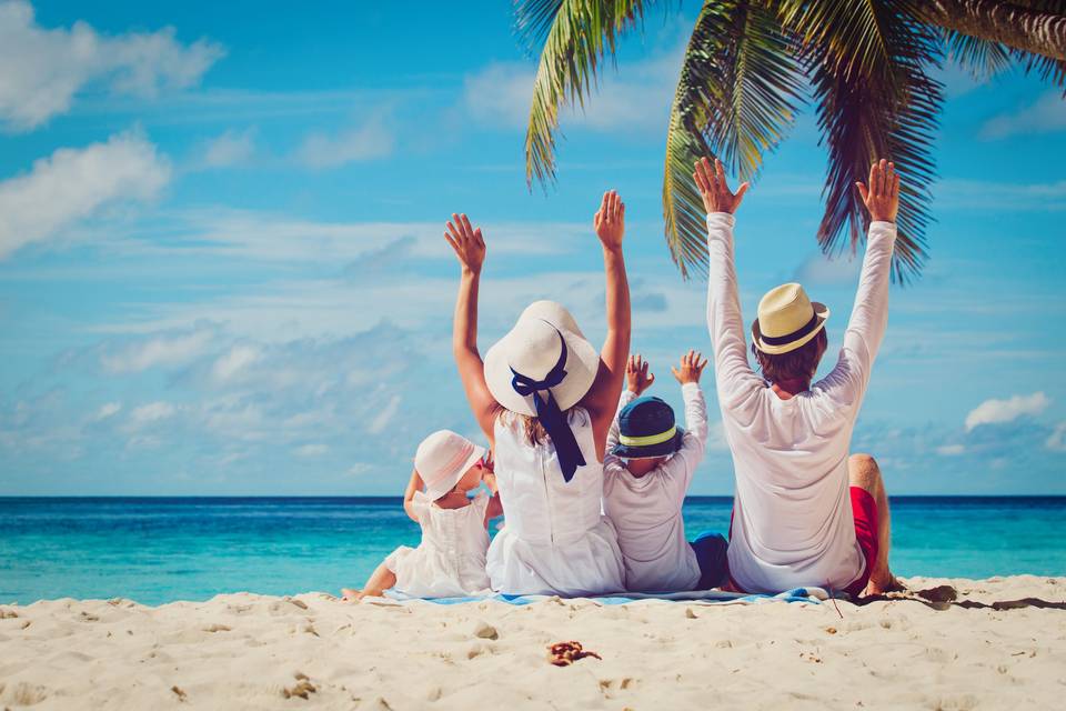 Hombre, mujer y dos niños pequeños, todos de espalda y con gorro, sentados en una playa bajo una palmera