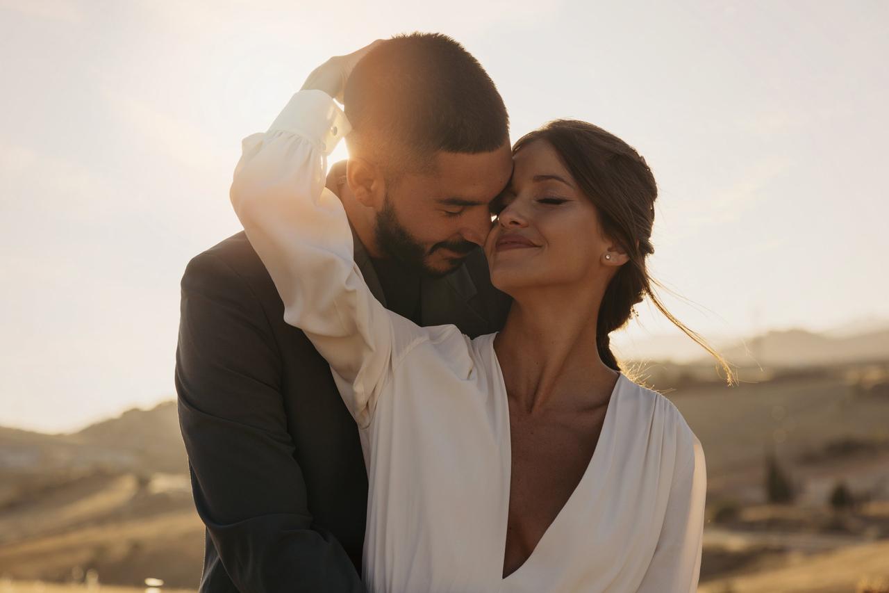 Mentiras de las mujeres: chico con traje y chica con blusa o vestido blanco de generoso escote en V se abrazan sonrientes durante una puesta de sol
