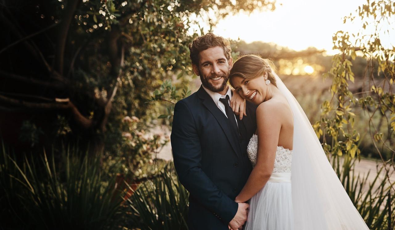 Cosas que las novias olvidan hacer el día de su boda: careja de recién casados muy sonrientes se abraza y coge de la mano en un ambiente al aire libre