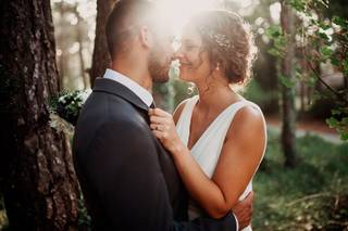 Chico con traje y chica con vestido blanco y el pelo recogido en un moño desenfadado con tocado juntan la nariz en un bonito exterior rodeados de naturaleza