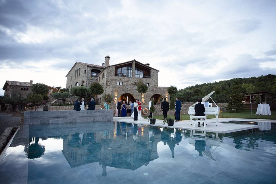 Música cóctel boda: bonito jardín con varias casas al fondo, una piscina en primer plano, un piano blanco y gente elegantemente vestida
