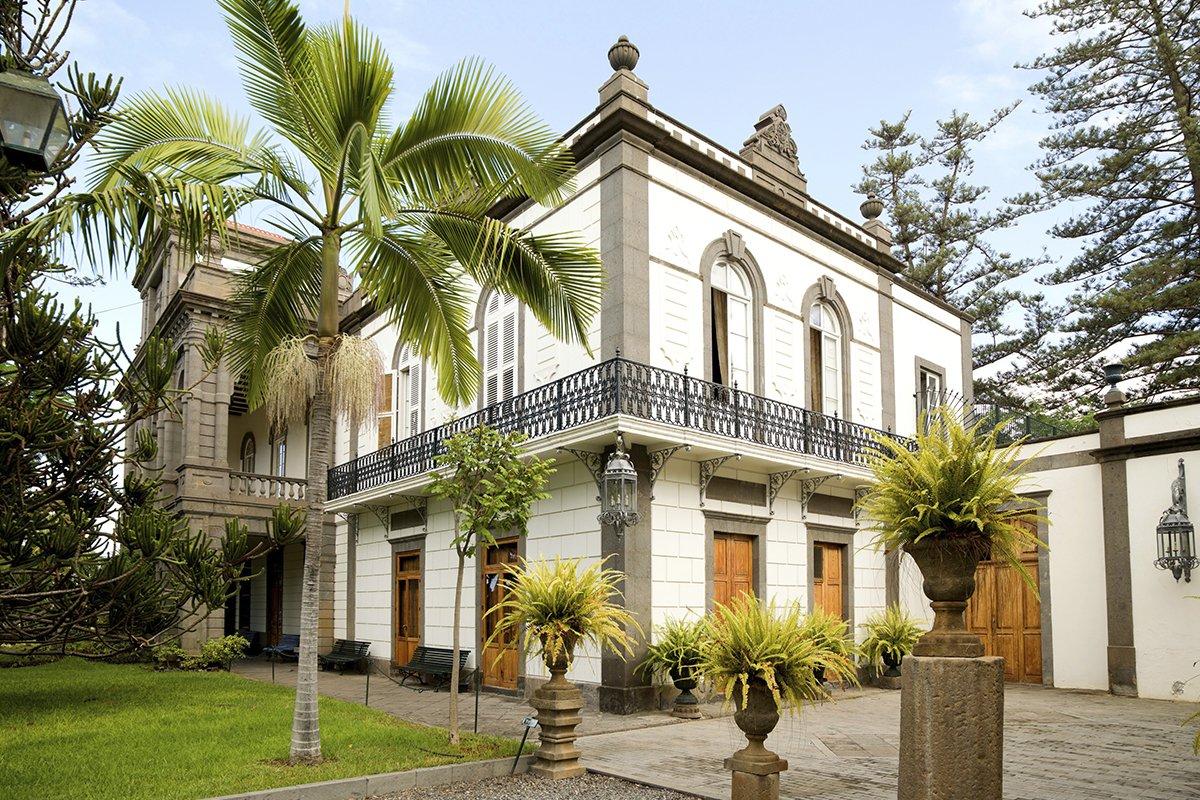 Fachada y jardín de una casa pintada en tonos claros, con cesped y palmeras