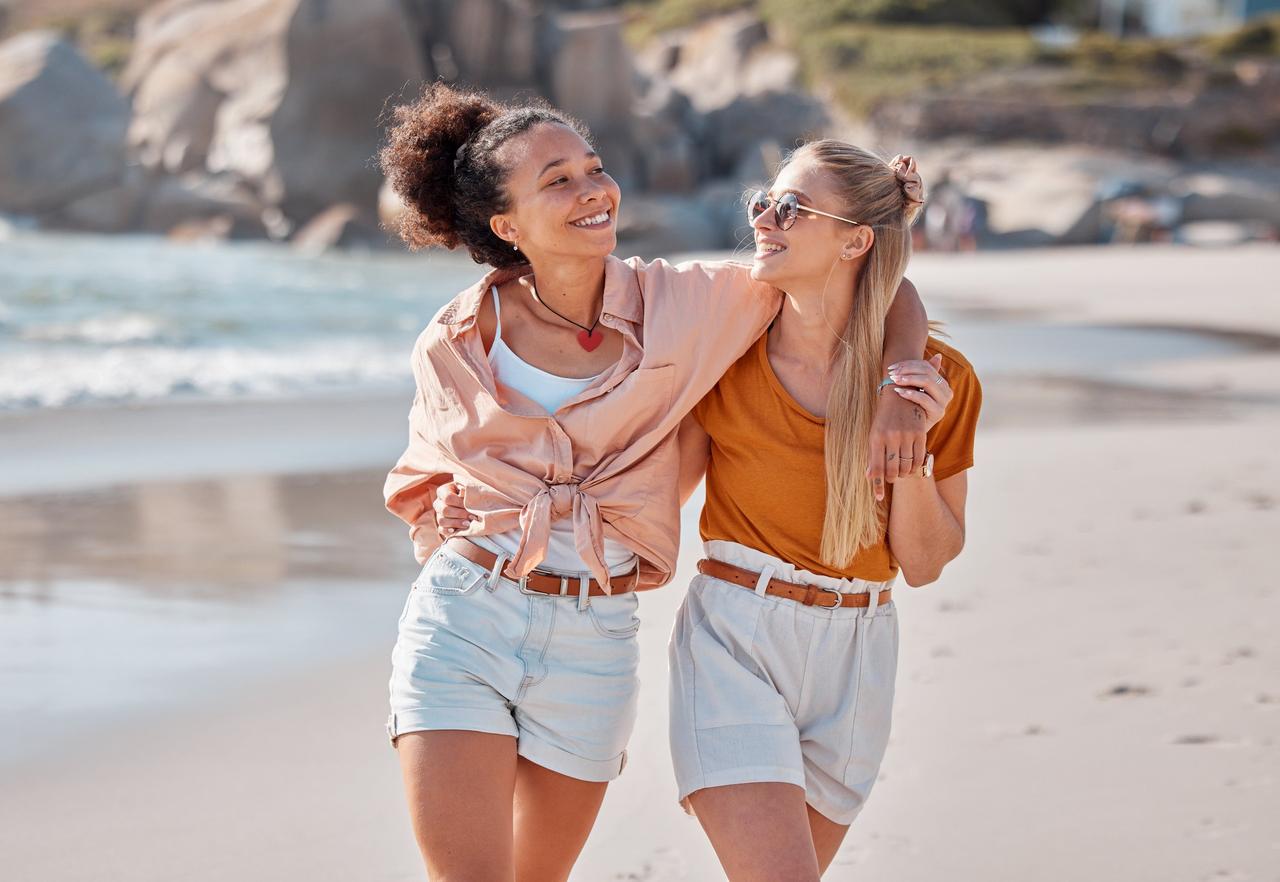 Señales de infidelidad: dos chicas jóvenes y sonrientes caminan abrazadas por la playa