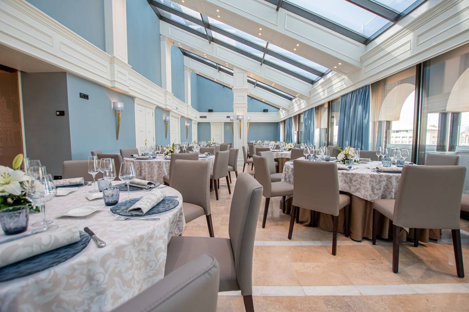 Espacioso salón con las paredes azules, grandes ventanales y mesas redondas, elegantemente vestidas para una comida o cena