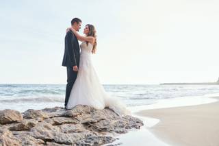 Chico con traje oscuro y chica con vestido de novia se abrazan sobre unas rocas junto al mar