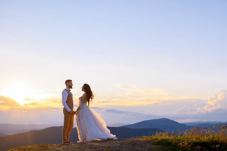 Chico con traje y pajarita y chica vestida de novia se cogen de la mano y se miran fijamente a los ojos en un precioso ambiente natural durante la puesta de sol