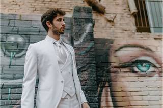 Chico con un traje blanco, con camisa y corbata en el mismo color junto a una pared exterior con un grafiti