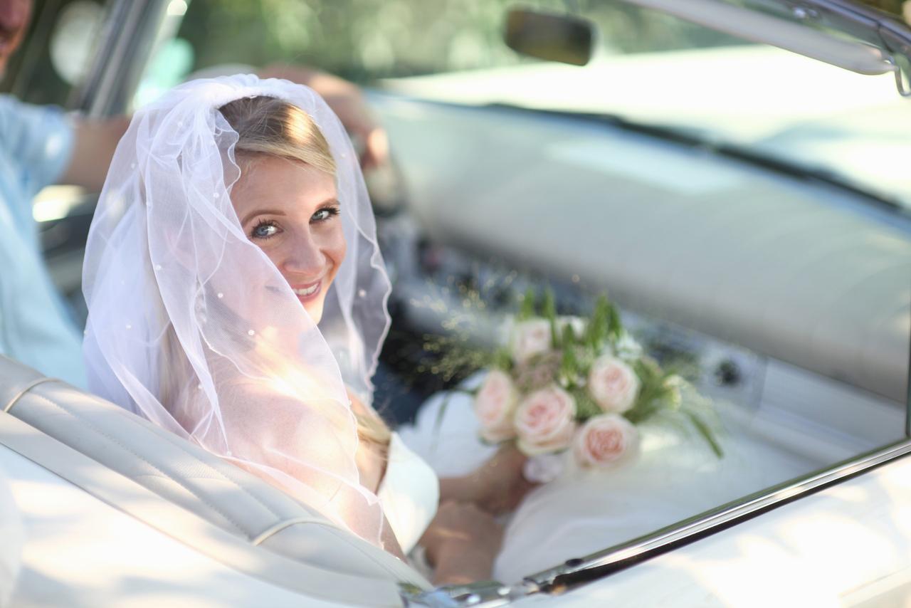 Cosas que las novias olvidan hacer el día de su boda: chica vestida de novia muy sonriente sentada en un coche descapotable con el ramo de flores en la mano