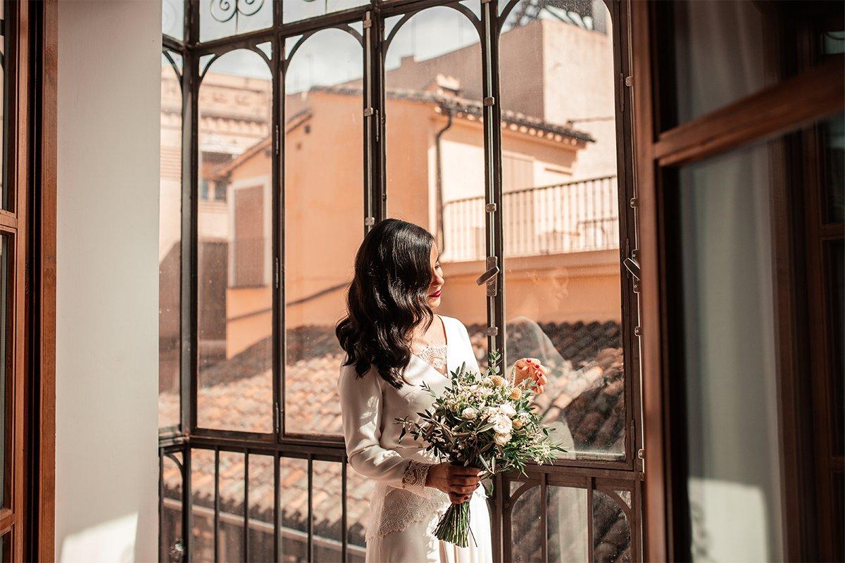 Chica con un vestido blanco, un ramo de flores en la mano y la melena suelta ondulada posa junto a un ventanal